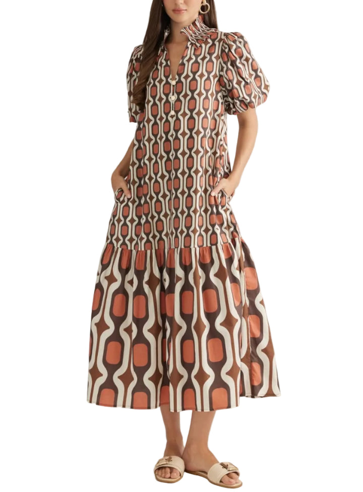 A woman modeling a midi dress with puff sleeves and a high neck, featuring a retro geometric pattern in rust orange, brown, and cream. She is standing against a plain light beige studio background.