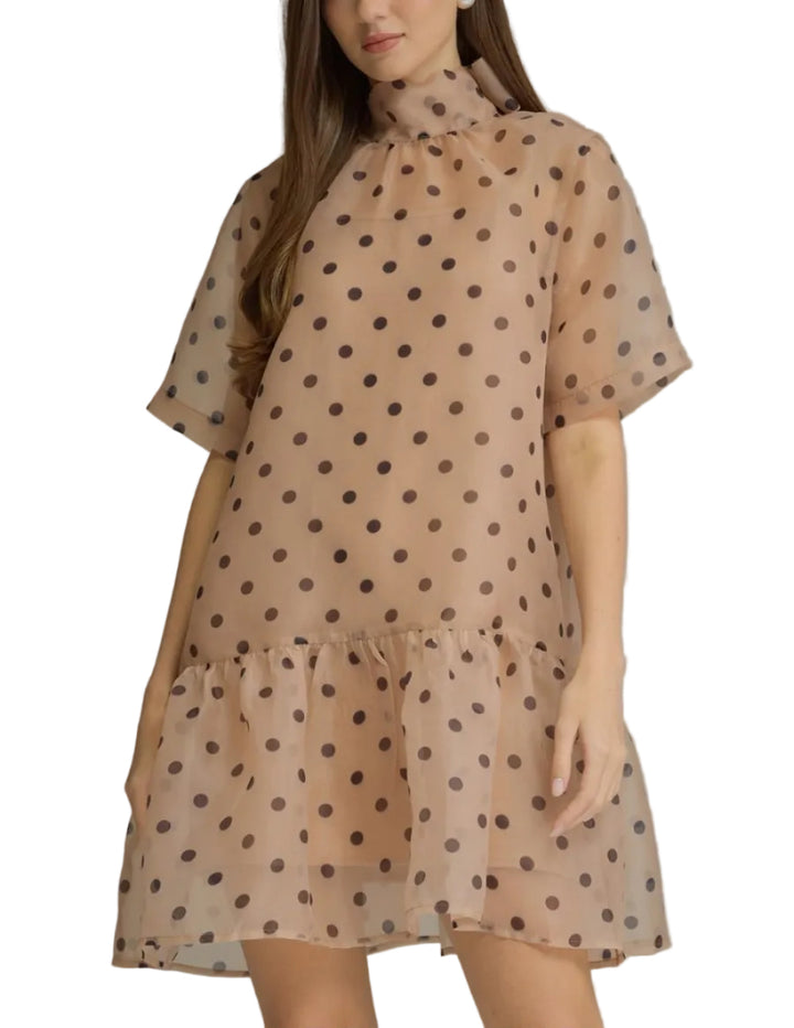 A woman modeling a sheer beige mini dress with dark brown polka dots, featuring a high mock neck and tiered ruffle hem, set against a plain gray studio background.