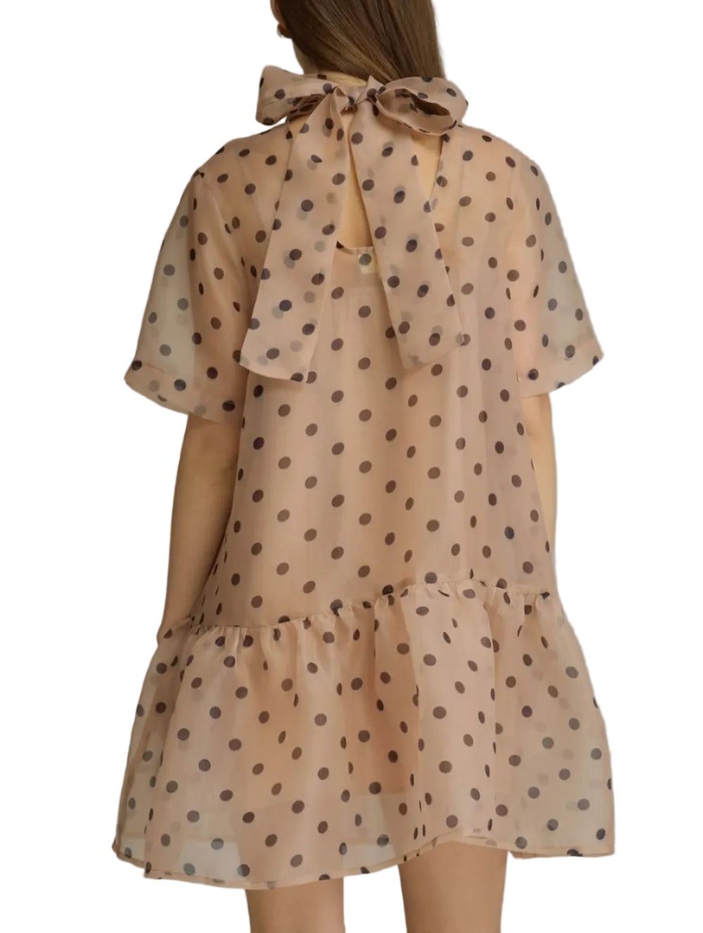 A woman viewed from the back wearing a sheer beige dress with dark brown polka dots and a large bow at the neckline, photographed against a plain light gray studio background.