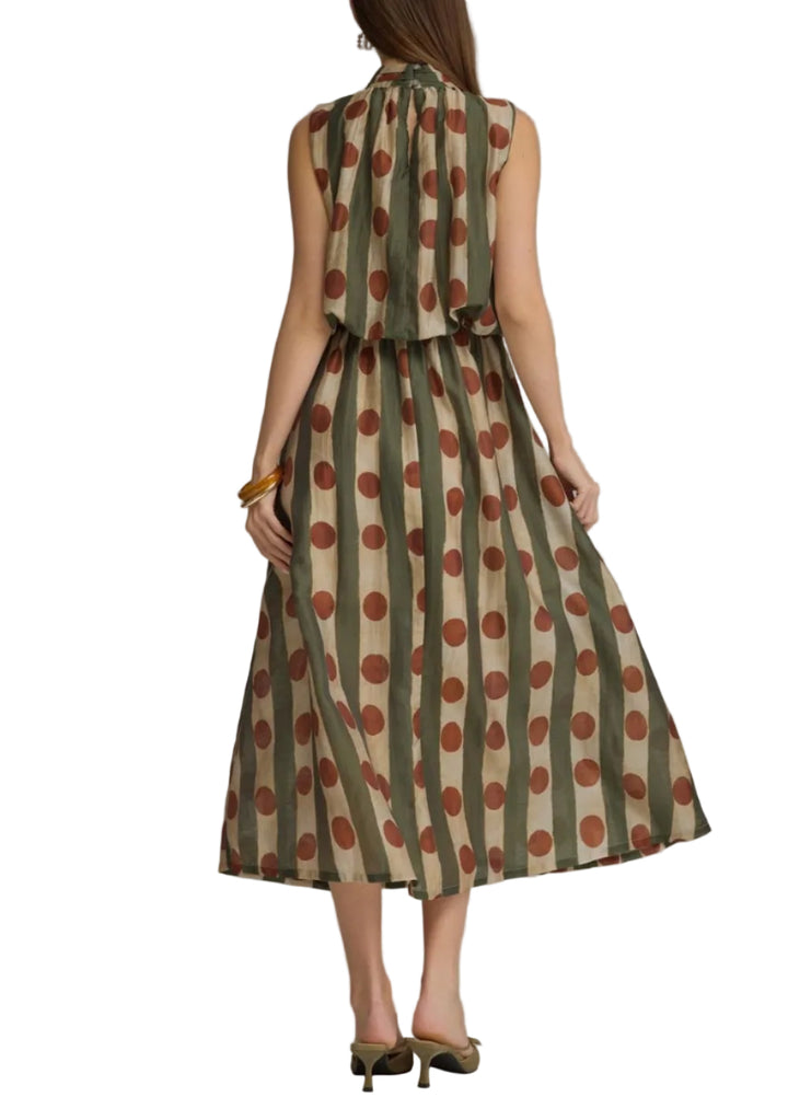 A woman viewed from the back wearing a sleeveless midi dress with vertical olive green and beige stripes and large rust-brown polka dots, paired with olive heels, against a neutral gray studio background.