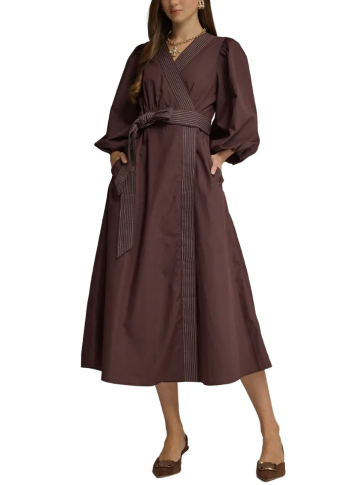 A woman models a dark brown midi wrap dress with puffed three-quarter sleeves, a matching waist tie belt, and vertical contrast stitching details on the front and sleeves, set against a solid gray studio background.