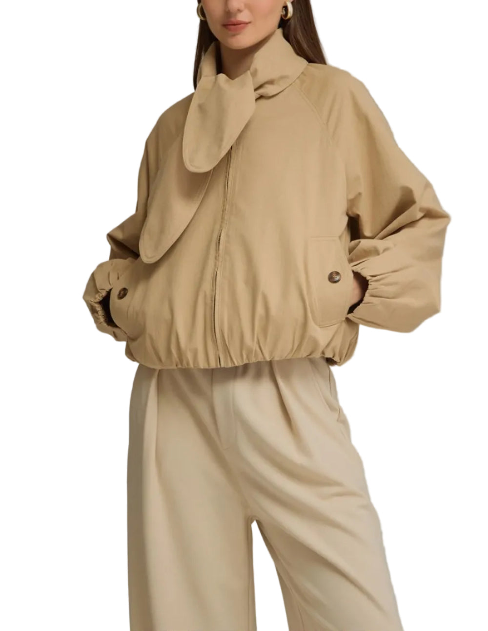 A woman modeling a beige cropped jacket with a large draped collar and matching beige wide-leg trousers, photographed against a plain light gray studio background.