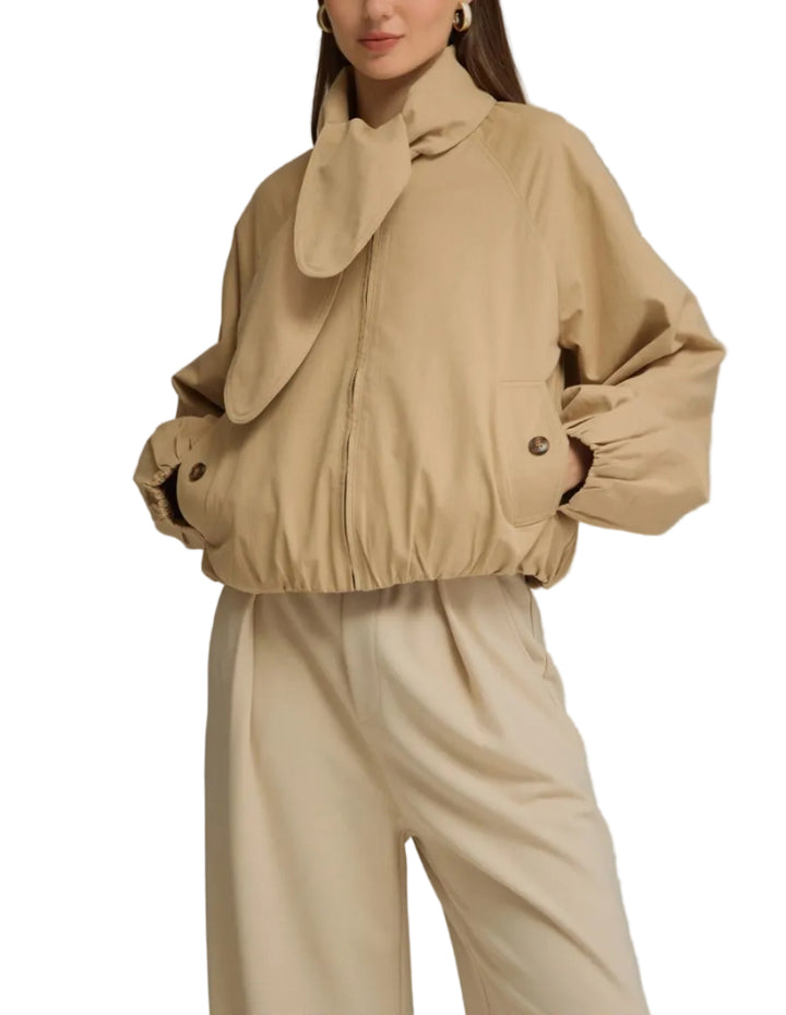 A woman modeling a beige cropped jacket with a large draped collar and matching beige wide-leg trousers, photographed against a plain light gray studio background.