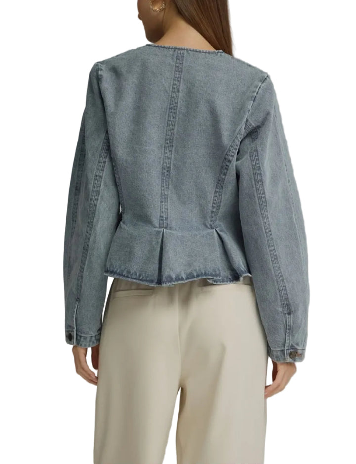 A woman shown from the back wearing a light blue denim jacket with a peplum waist and cream-colored trousers, set against a solid gray studio background.