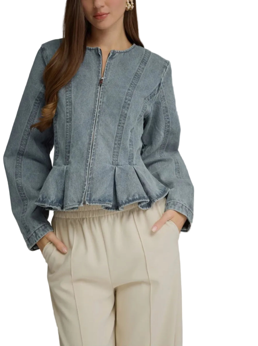 A woman wearing a cropped light blue denim jacket with a front zipper and peplum hem, paired with cream-colored trousers, against a solid gray studio background.