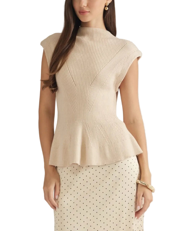 A woman modeling a ribbed knit sleeveless top with a high mock neck and peplum hem in a light cream color, paired with a cream polka dot skirt against a solid light gray background.