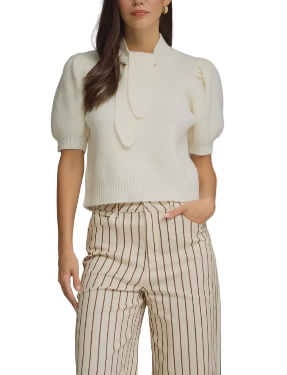A woman modeling a cream short-sleeve knit top with a large bow detail at the neckline, paired with beige and brown vertically striped trousers, against a solid gray studio background.