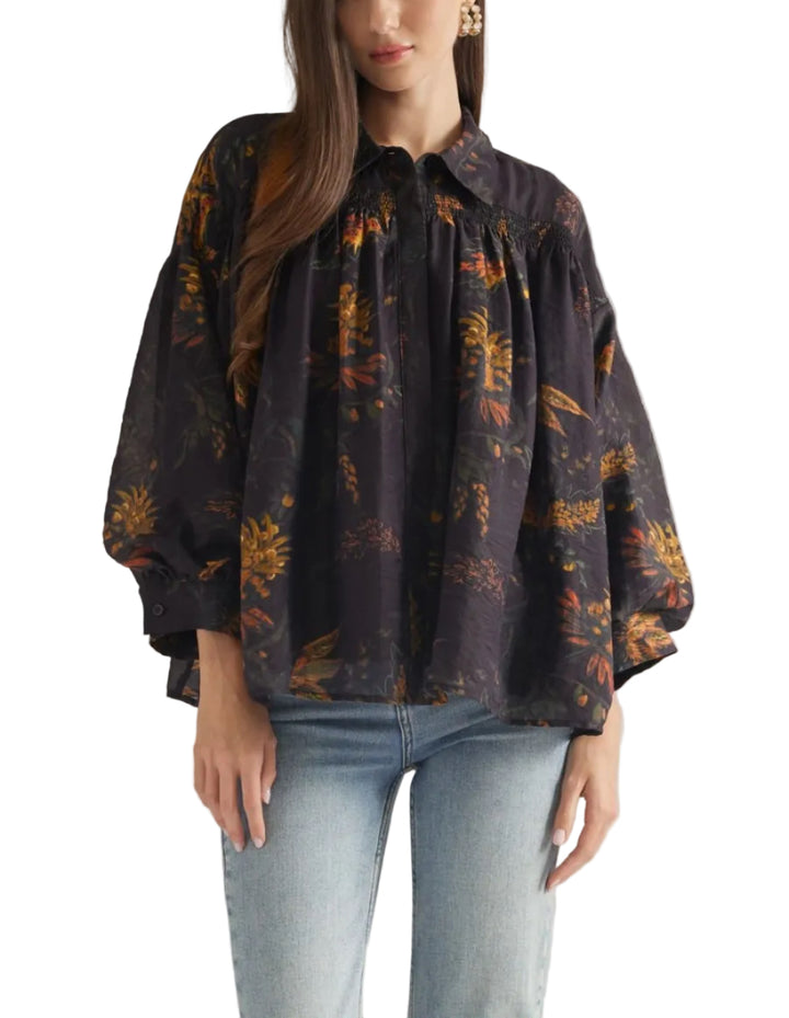 A woman modeling a dark, loose-fitting blouse with a tropical floral print in orange and yellow, paired with light-wash blue jeans, against a solid light gray background.