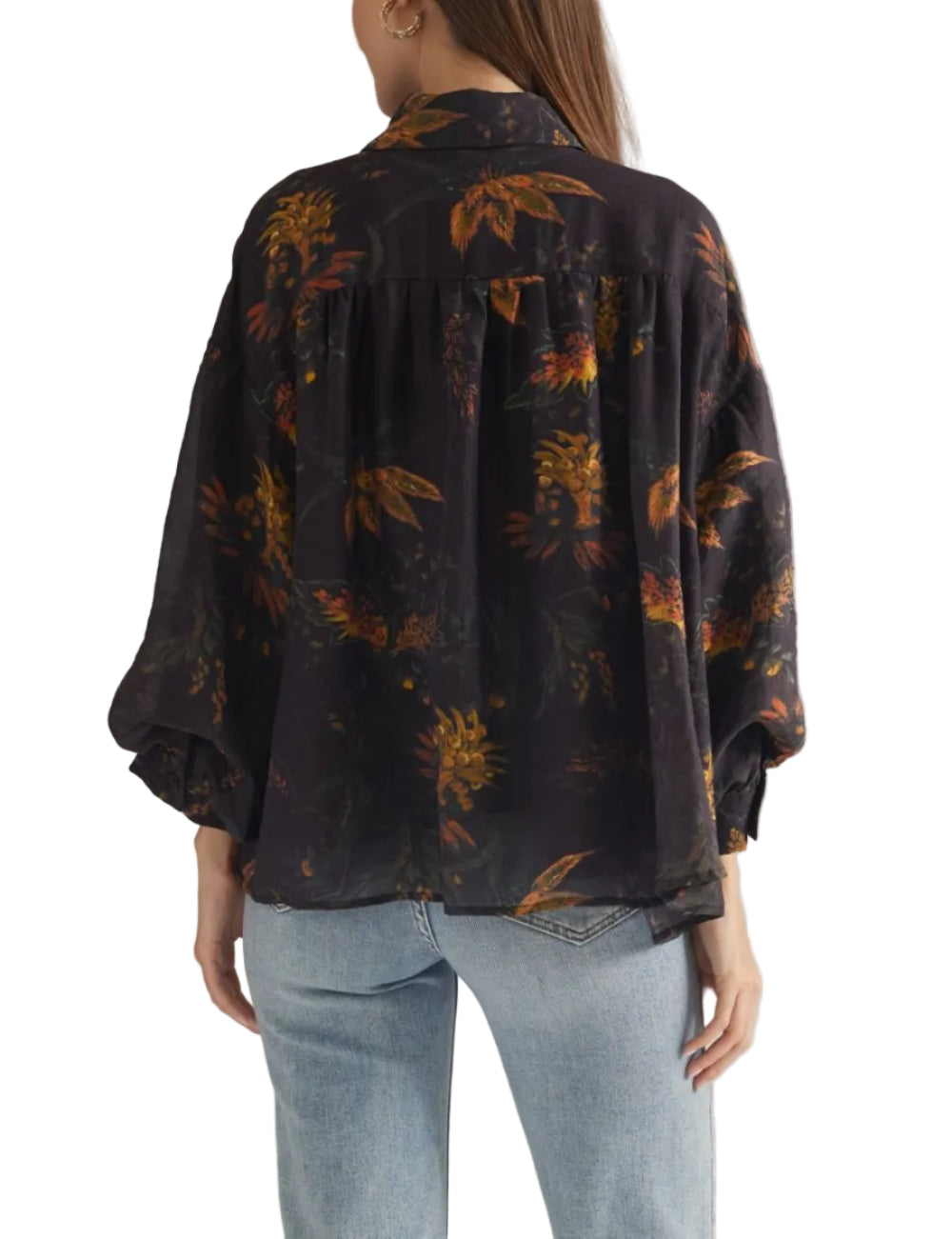A woman shown from the back wearing a dark floral blouse with voluminous sleeves and light-wash jeans, photographed against a plain light gray studio background.