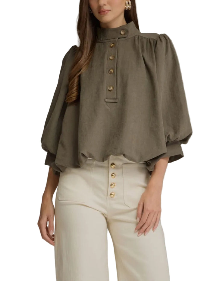A woman modeling an olive green puff-sleeve blouse with gold-tone buttons down the front, paired with cream-colored high-waisted trousers, against a solid gray studio background.