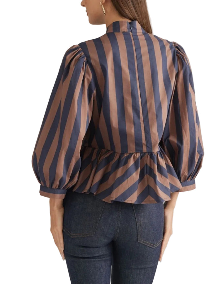 A woman shown from the back wearing a high-neck blouse with vertical brown and navy stripes, puffed three-quarter sleeves, and a peplum waist, paired with dark blue jeans against a plain beige studio background.