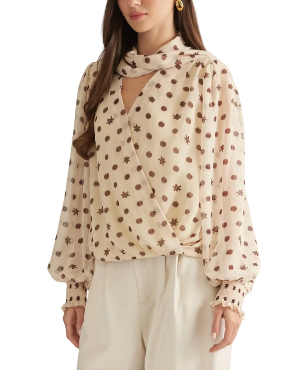 A woman modeling a beige long-sleeve blouse with brown polka dots and star motifs, featuring a keyhole cutout at the chest and a matching neck scarf, paired with cream trousers against a light gray studio background.