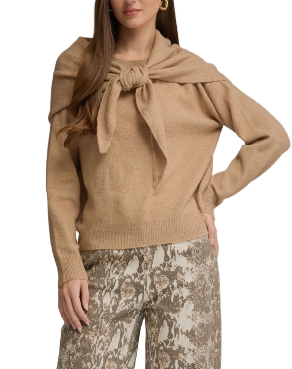 A woman modeling a camel-colored knit sweater with a large bow detail at the neckline, paired with patterned pants, against a solid gray studio background.