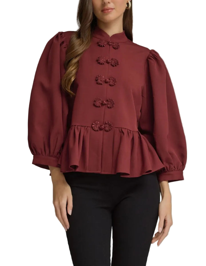 A woman modeling a burgundy long-sleeve blouse with puff sleeves, a high collar, and decorative button details, paired with black pants against a neutral gray studio background.