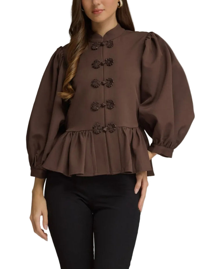 A woman modeling a brown long-sleeve blouse with puff sleeves, a high collar, and decorative button details down the front, paired with black pants against a neutral gray studio background.