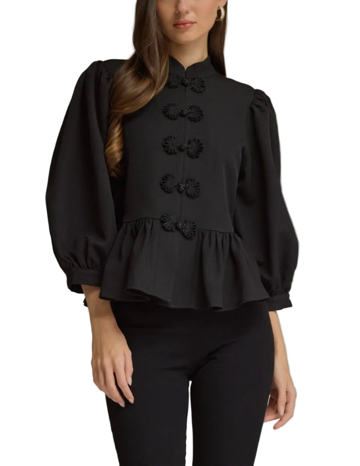 A woman modeling a black long-sleeve blouse with a high mock neck, voluminous puff sleeves, and a peplum hem, featuring decorative black fabric flowers down the front. She is wearing black pants against a solid gray studio background.