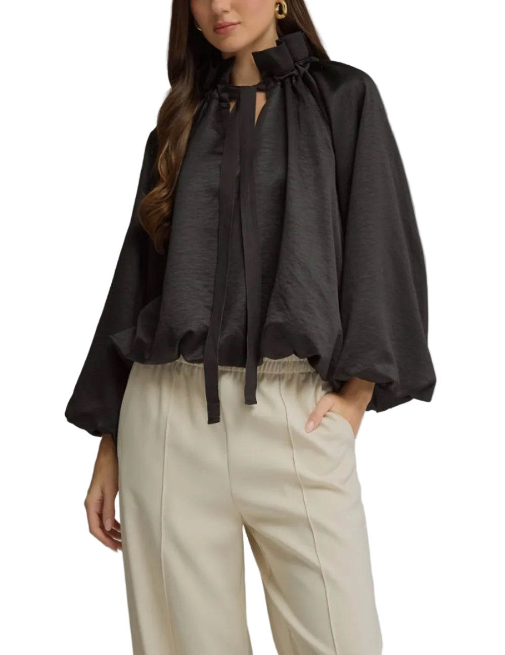 A woman modeling a black long-sleeve blouse with a high gathered neckline and front tie detail, paired with cream-colored trousers, against a solid gray studio background.