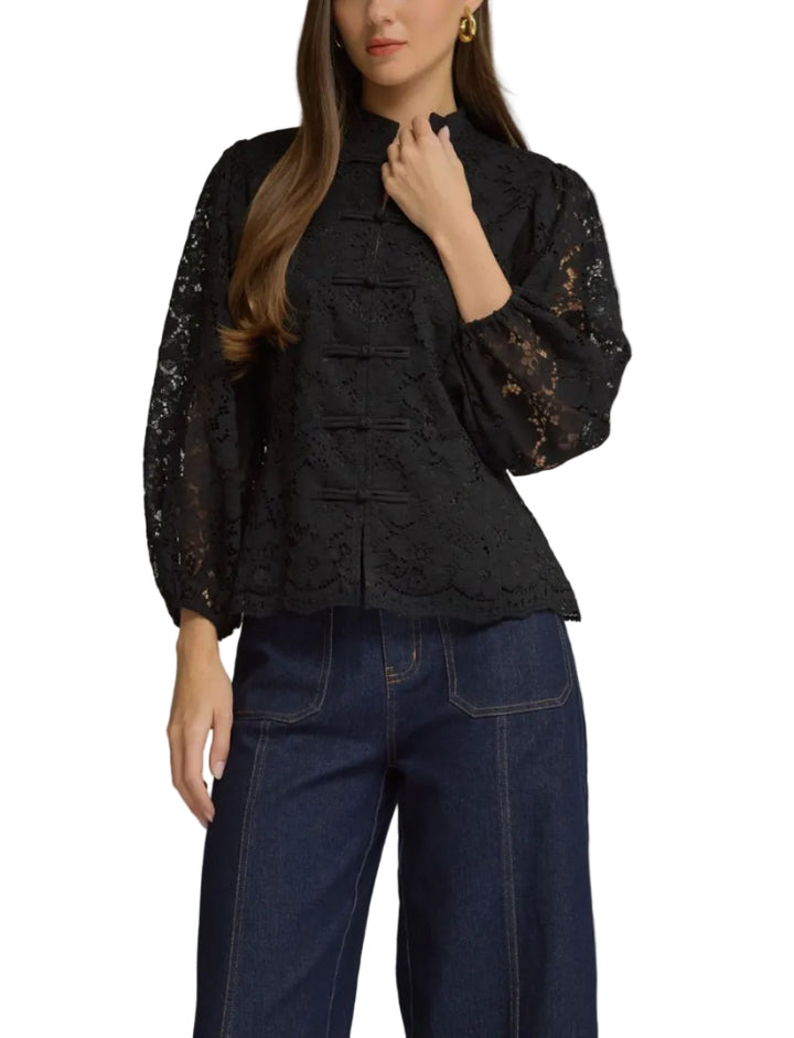A woman modeling a black lace long-sleeve blouse with a high mock neck and decorative front buttons, paired with dark blue jeans, against a solid gray studio background.