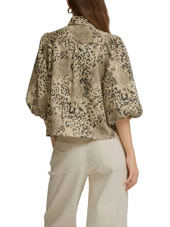 A woman shown from the back wearing a cropped leopard-print blouse with voluminous puff sleeves and light beige trousers, photographed against a plain gray studio background.