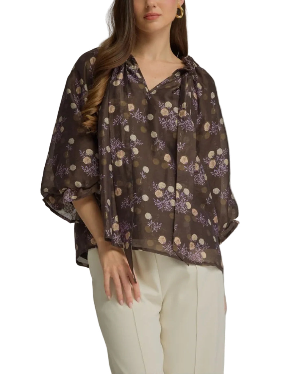 A woman modeling a sheer brown blouse with a purple and beige floral pattern, paired with cream trousers, against a solid gray studio background.