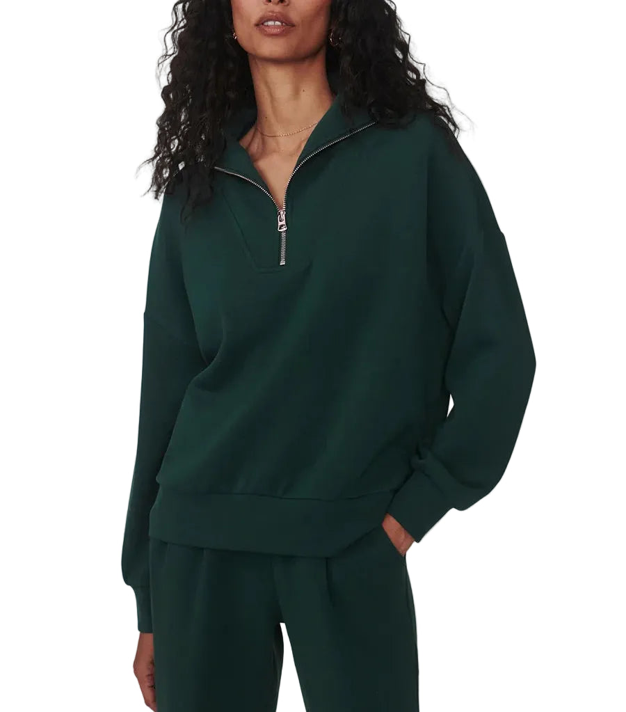 A woman modeling a dark green half-zip sweatshirt and matching sweatpants against a light gray studio background.