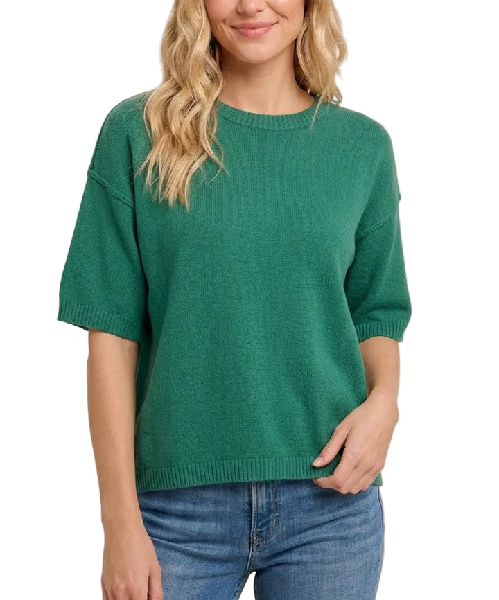 A woman wearing a green short-sleeve knit sweater and blue jeans, standing against a plain light beige background.