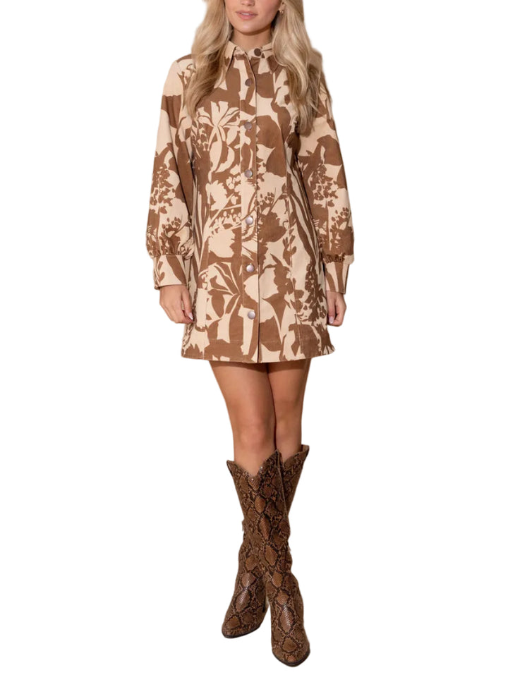 A woman modeling a long-sleeve mini dress with a brown and beige botanical pattern, paired with brown snakeskin-patterned cowboy boots, against a solid beige studio background.