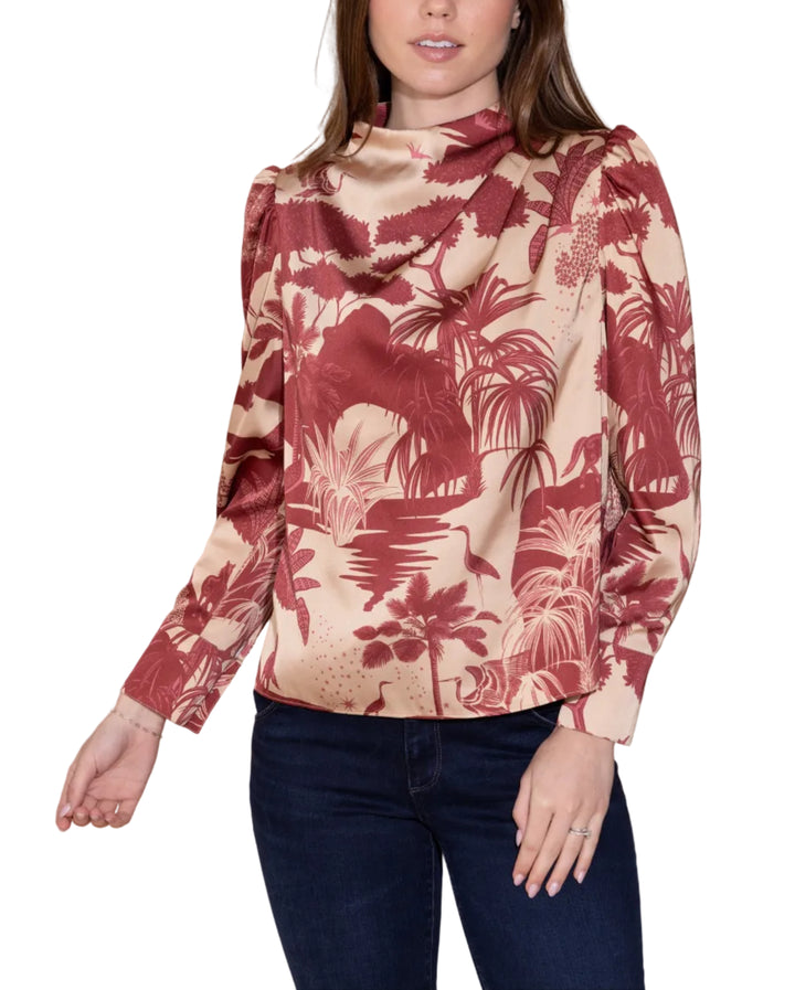 A woman modeling a long-sleeve satin blouse with a tropical leaf print in deep red and beige, paired with dark blue jeans, against a solid beige studio background.