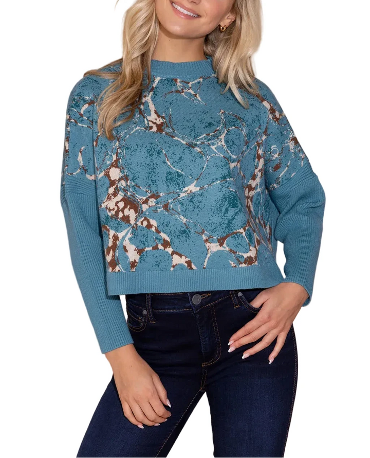 A woman wearing a teal blue knit sweater with a brown and white abstract animal print pattern, paired with dark blue jeans, standing against a solid beige background.