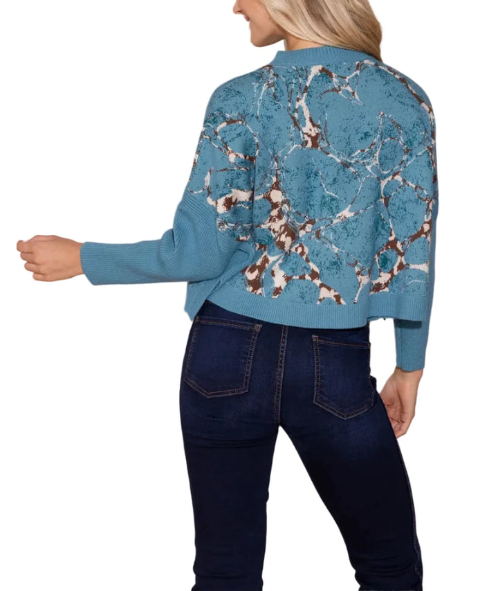 A woman shown from the back wearing a cropped blue knit sweater with a brown and white marble pattern, paired with dark blue jeans, against a solid beige background.