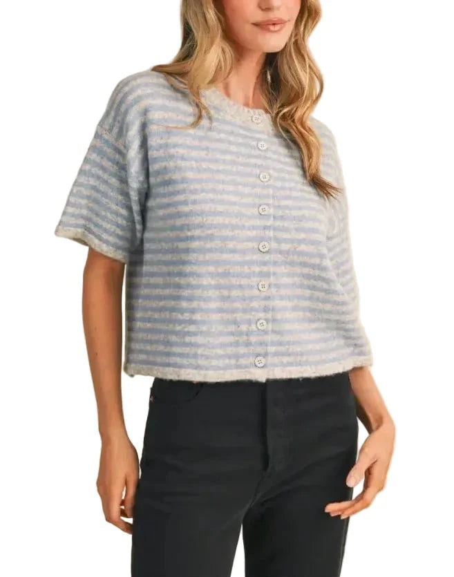 A woman wearing a light blue and white striped short-sleeve cropped button-up top with black pants, standing against a solid beige background.