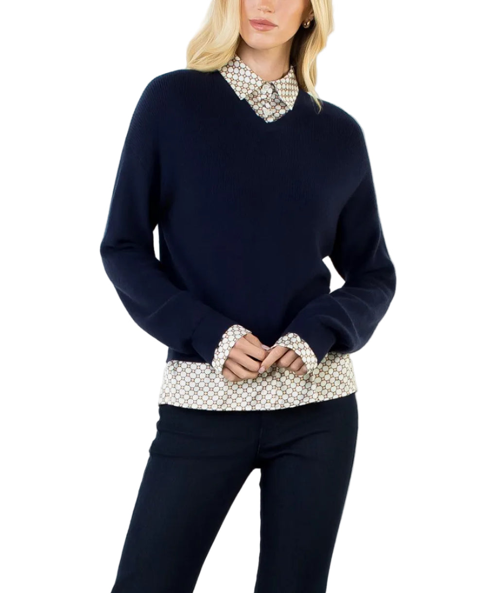 A blonde woman modeling a navy blue V-neck sweater layered over a beige and black patterned collared shirt, paired with dark navy pants, against a plain white studio background.