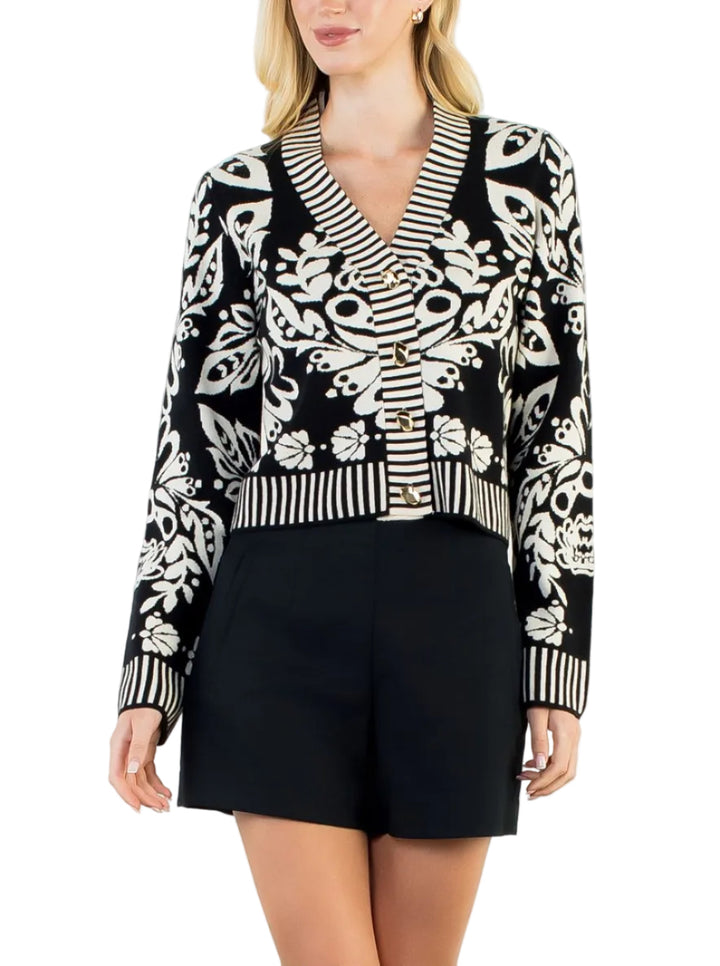 A woman modeling a black and white patterned knit cardigan with striped trim, paired with black shorts, against a plain white studio background.